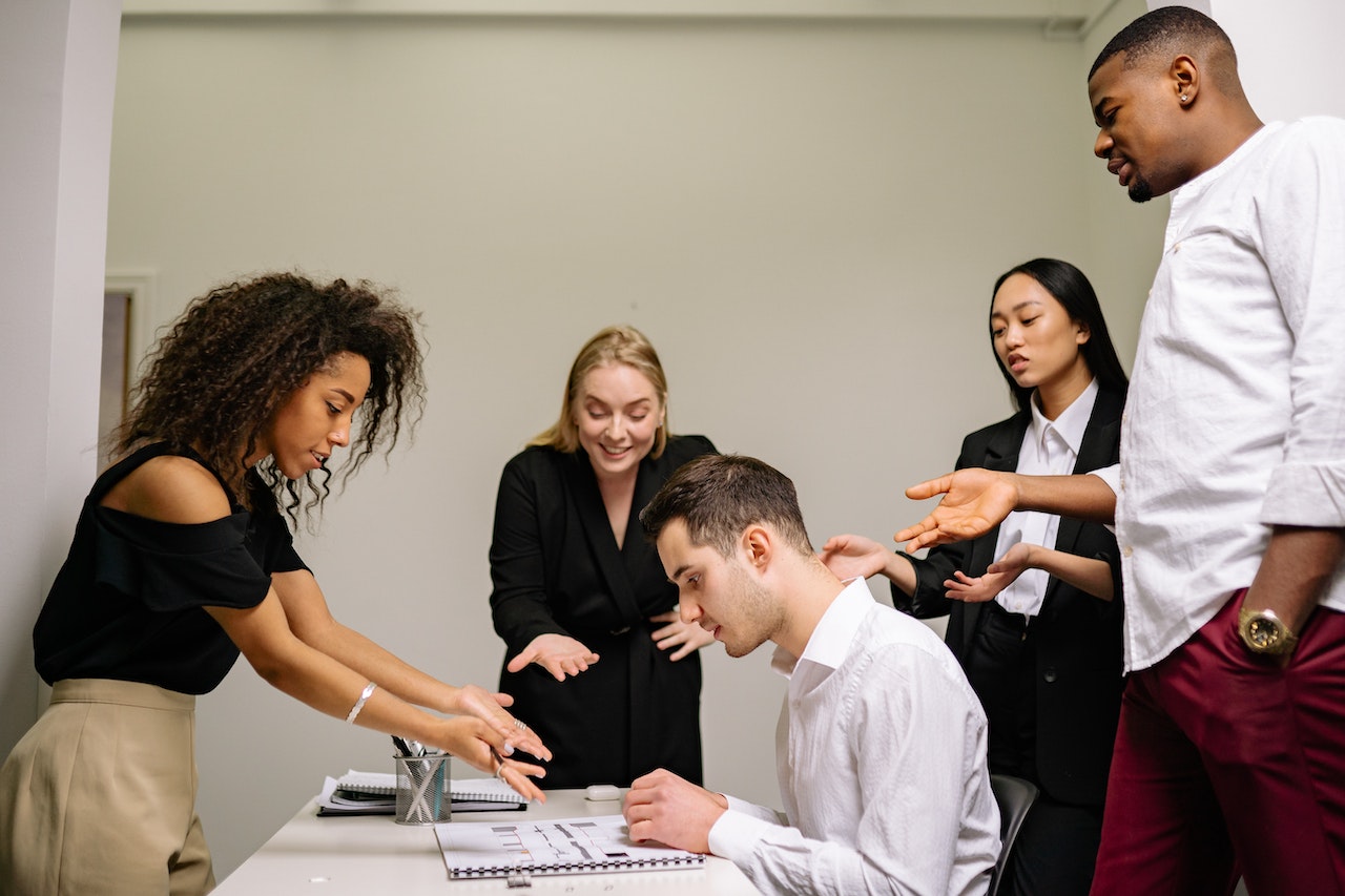 Multiple People Standing Around a Desk and Pointing at the Person Working There